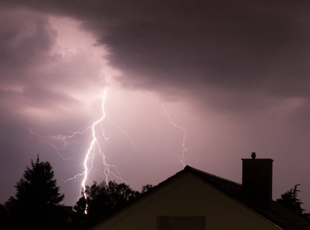 how to file a roof insurance claim bolt of lightning against purple and gray sky with silhouette of trees and houses in the foreground