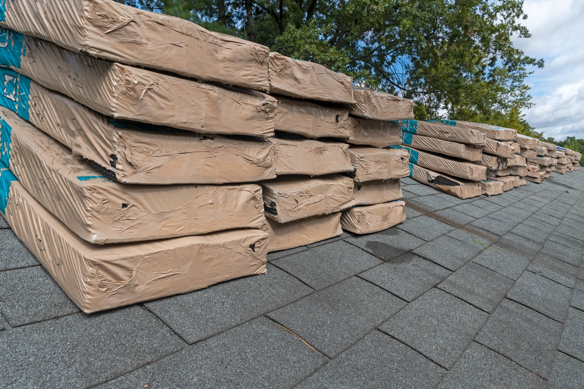 Several stacks of packaged asphalt shingles await installation on a rooftop, framed by trees and a cloudy sky. Wrapped in brown and blue packaging, these shingles are ready for action, reminding homeowners to consider how often to replace their roof for optimal protection.