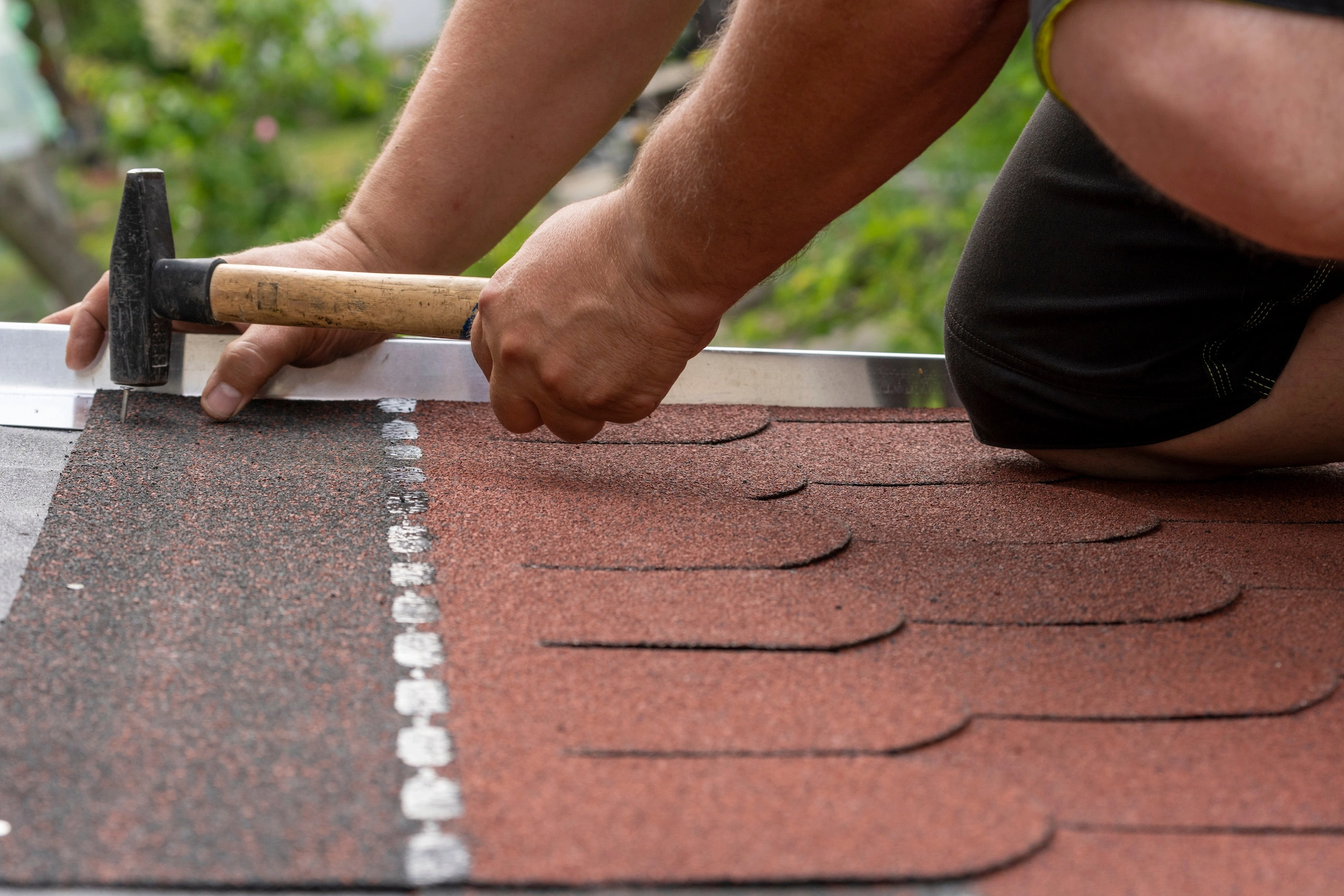A person skillfully engages in DIY roof repair, using a hammer to install red shingles on a rooftop. The focus is on the hands and tools, set against a backdrop of blurred greenery.