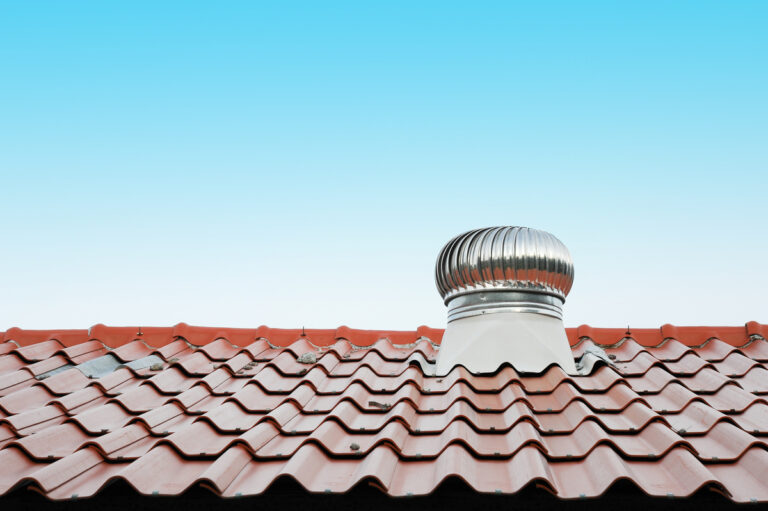 A red tile roof with a sleek silver metal turbine vent, one of the various types of roof vents, stands out against the clear blue sky.