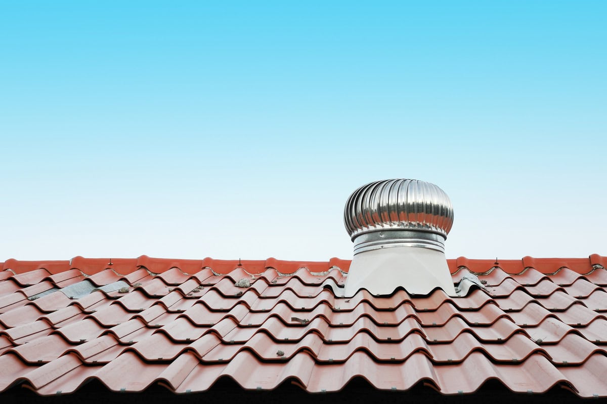 A red tile roof with a sleek silver metal turbine vent, one of the various types of roof vents, stands out against the clear blue sky.