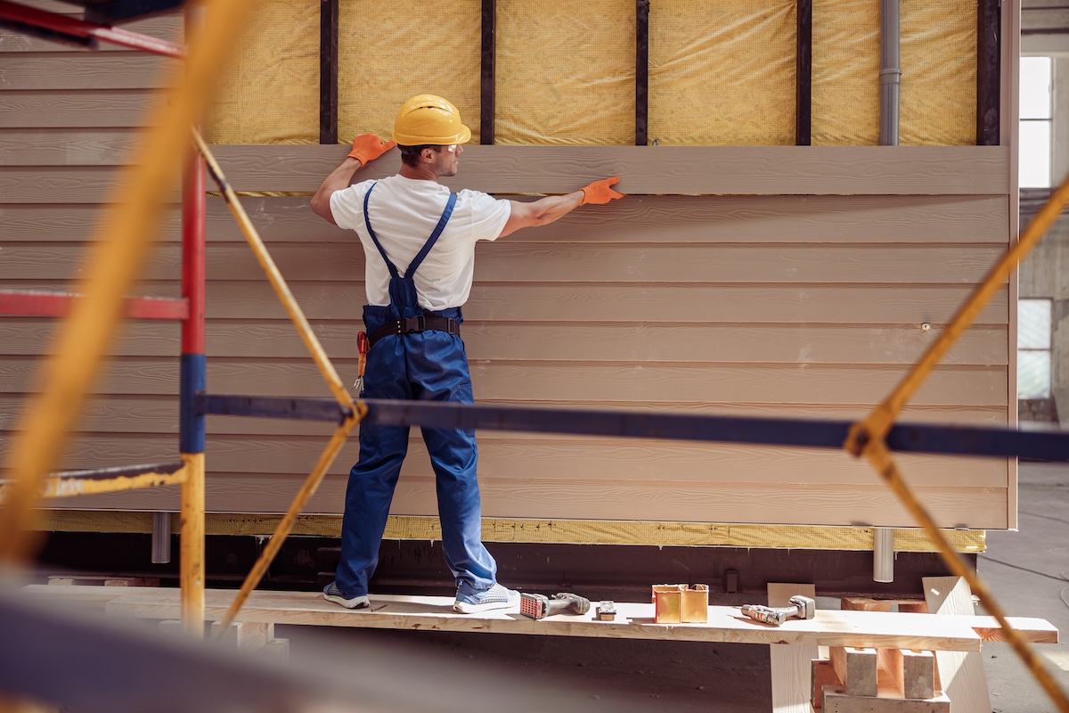 Back view of young man builder wearing safety helmet and work overalls while installing exterior wood siding