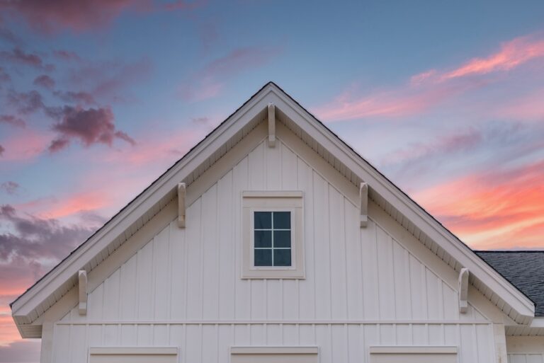New gable with decorative brackets, white vertical vinyl lap siding with square attic window on a newly built home in America colorful sunset background
