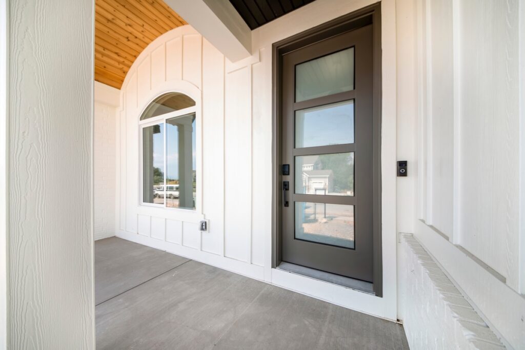 Exterior of a house with white board and batten sidings. Porch of a house with gray front door with glass panels and doorbell camera near the arched window.