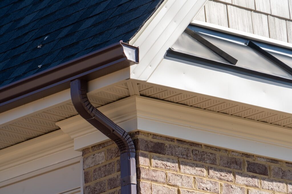 Closeup view of dark brown gutter system with white soffit vent, gutter guard, drop outlet, downspout,