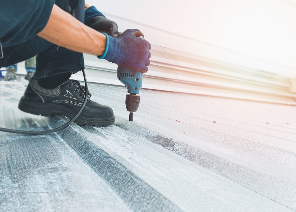 Workers wearing protective gear are screwing nuts on the metal roof with electric drills to install or repair with professional expertise, an orange light shines through.