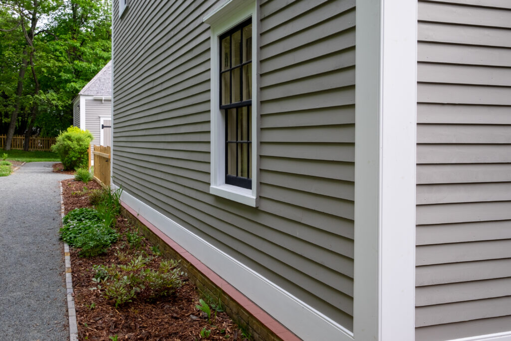 Old tan colored house with white trim and black wood window frame spacers. The glass in the window is wavy and reflects the sun. The exterior wall is made of vintage beige cape cod clapboard siding.